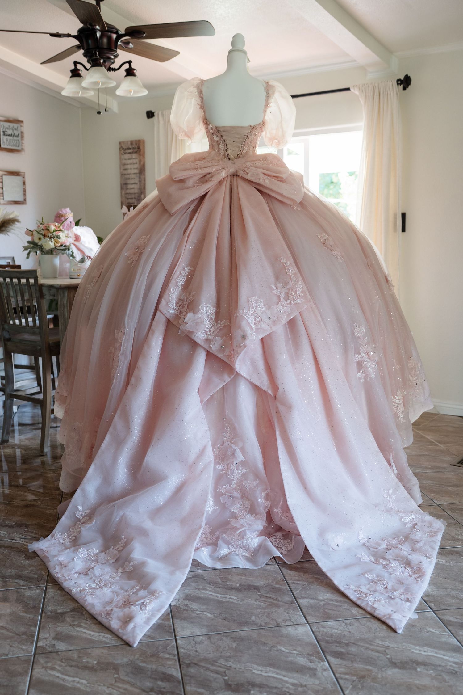 Elegant blush pink quinceañera ball gown with lace trim, large bow back, and layered skirt displayed on mannequin indoors.