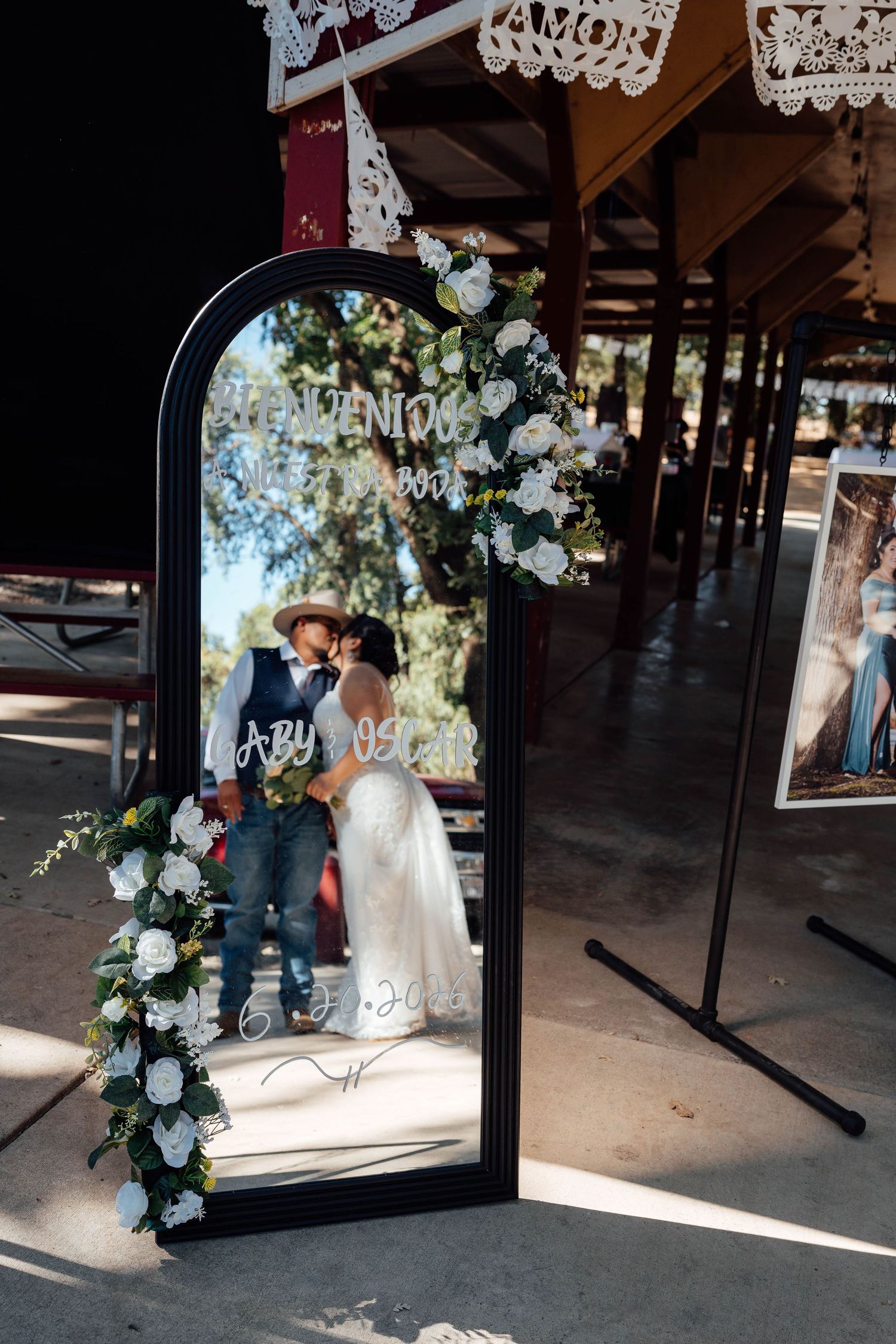 Newlyweds kiss reflected in decorative mirror with floral arch at reception.