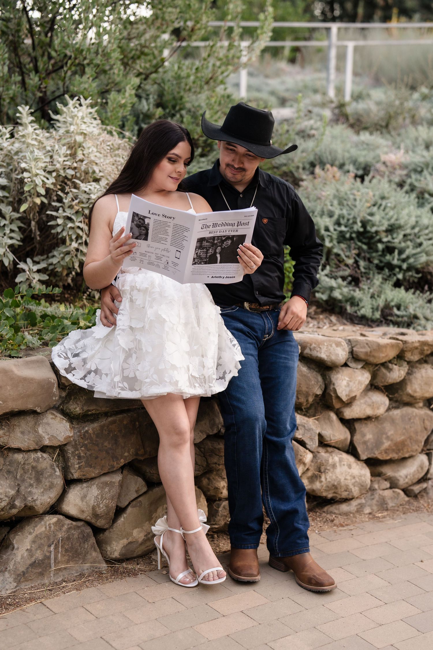 Couple reading a newspaper together on a stone wall, woman in white dress, man in black cowboy hat.