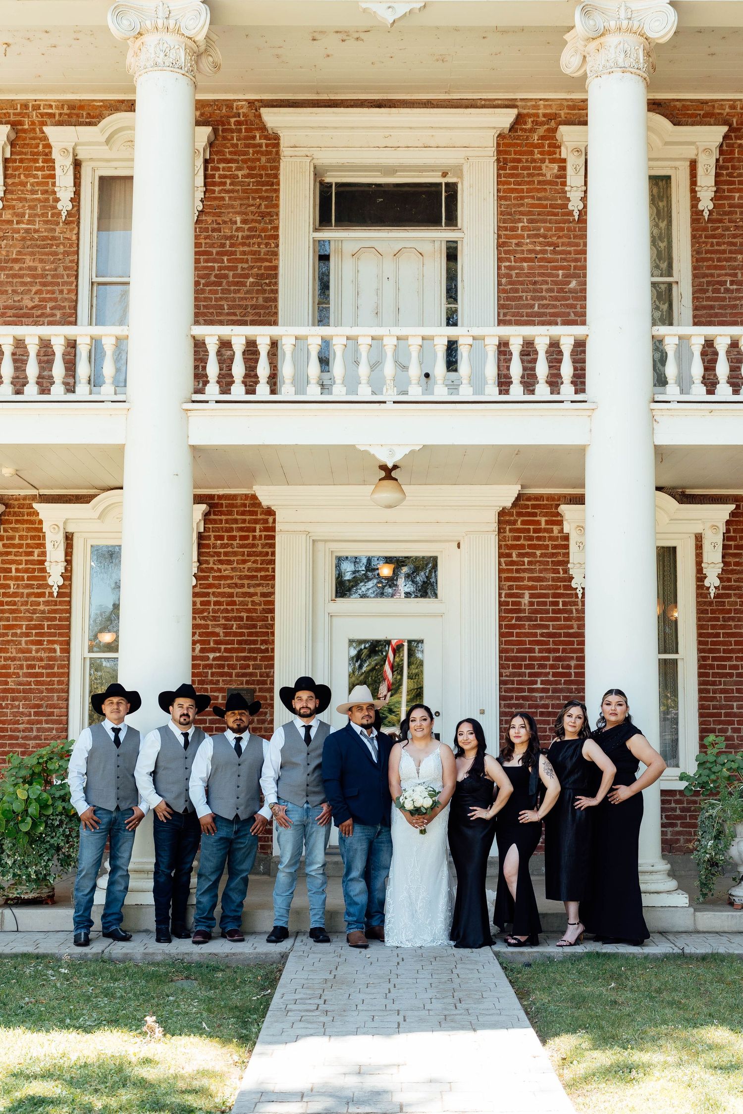 Wedding party poses in front of historic brick mansion with groomsmen in cowboy hats and bridesmaids in black.