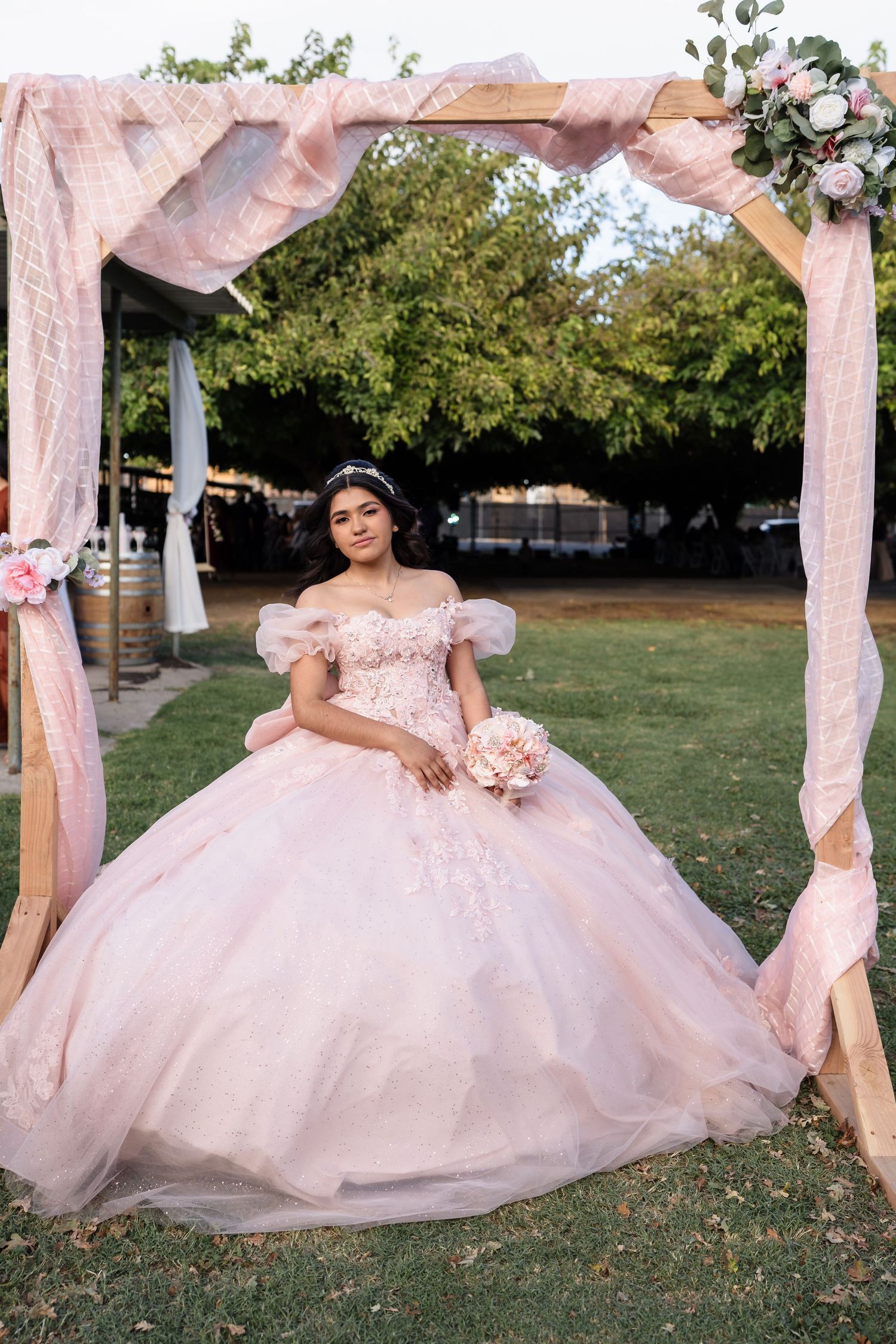 Teen in pink quinceañera ball gown holds bouquet under floral arch at outdoor celebration.