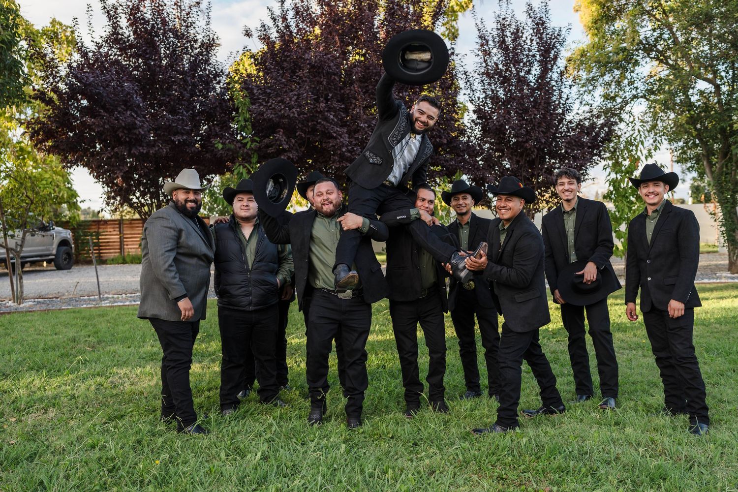 Group of men in black suits and cowboy hats posing outdoors, one lifted in the air, with red-leafed trees behind.
