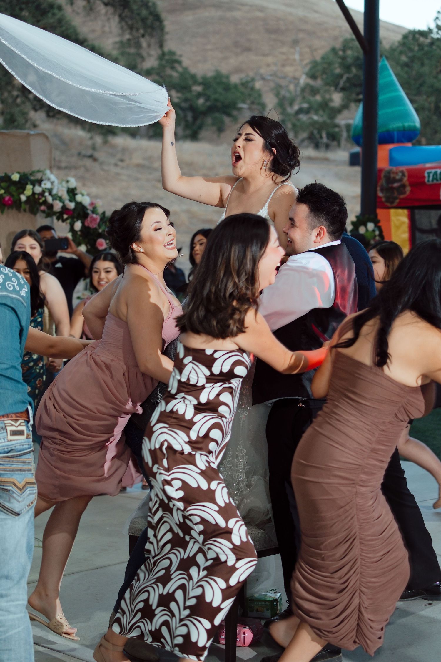 Guests dancing joyfully at an outdoor wedding reception, with a bride in a pink dress raising a veil.