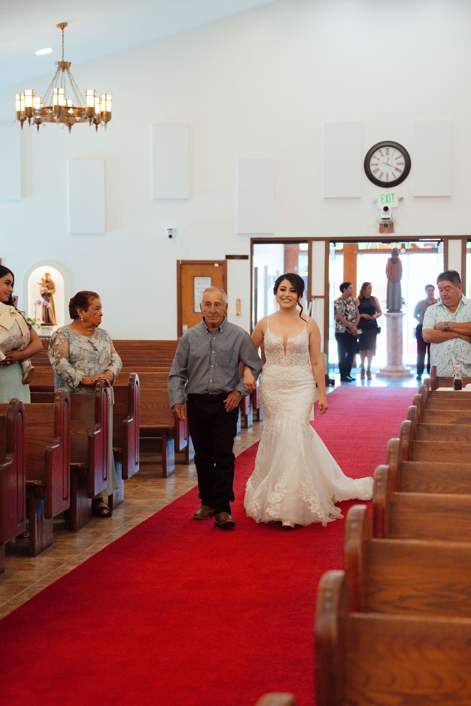 Bride in lace mermaid gown walks down red carpet aisle with escort in white church wedding ceremony.