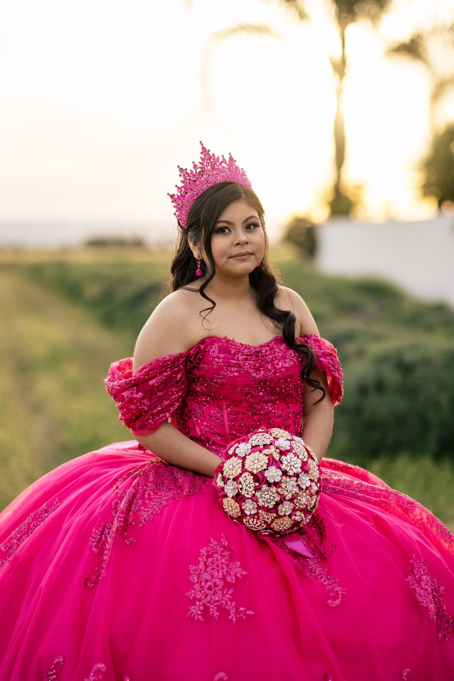 Teen girl in vibrant pink quinceañera dress with crown holds bouquet at golden hour outdoors.