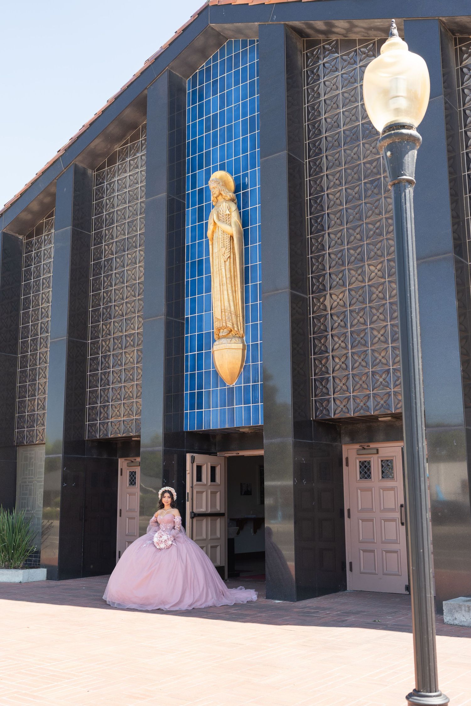 Quinceañera in pink ball gown stands before a modern Catholic church facade with golden statue.
