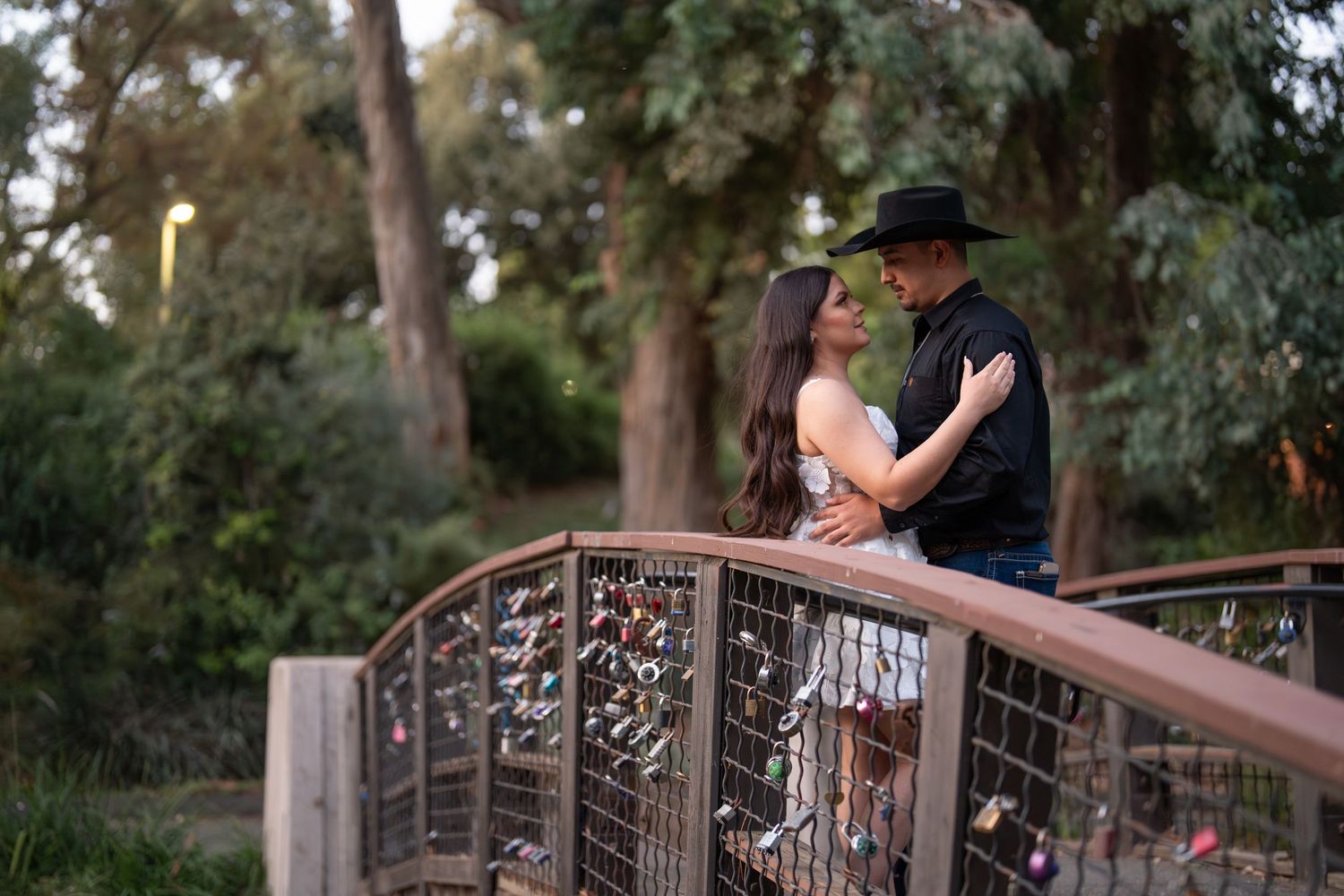 Couple embracing on a bridge decorated with love locks surrounded by lush green trees.
