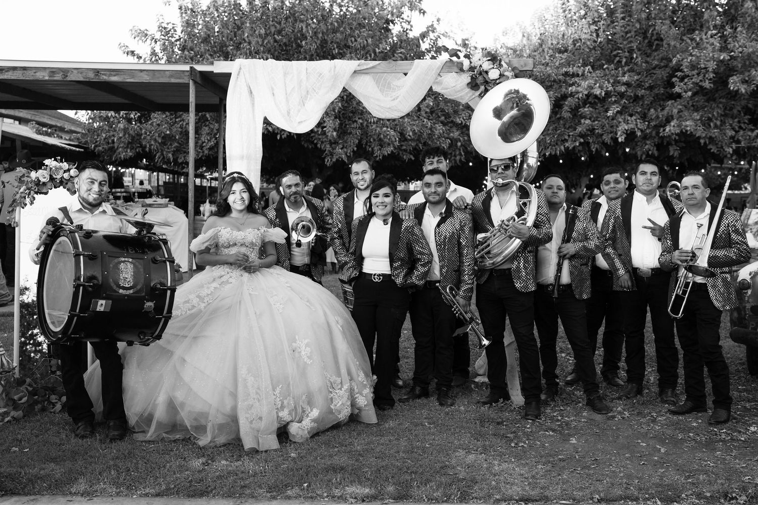 Black and white photo of a quinceañera girl in a ball gown posing with a brass band at an outdoor celebration.