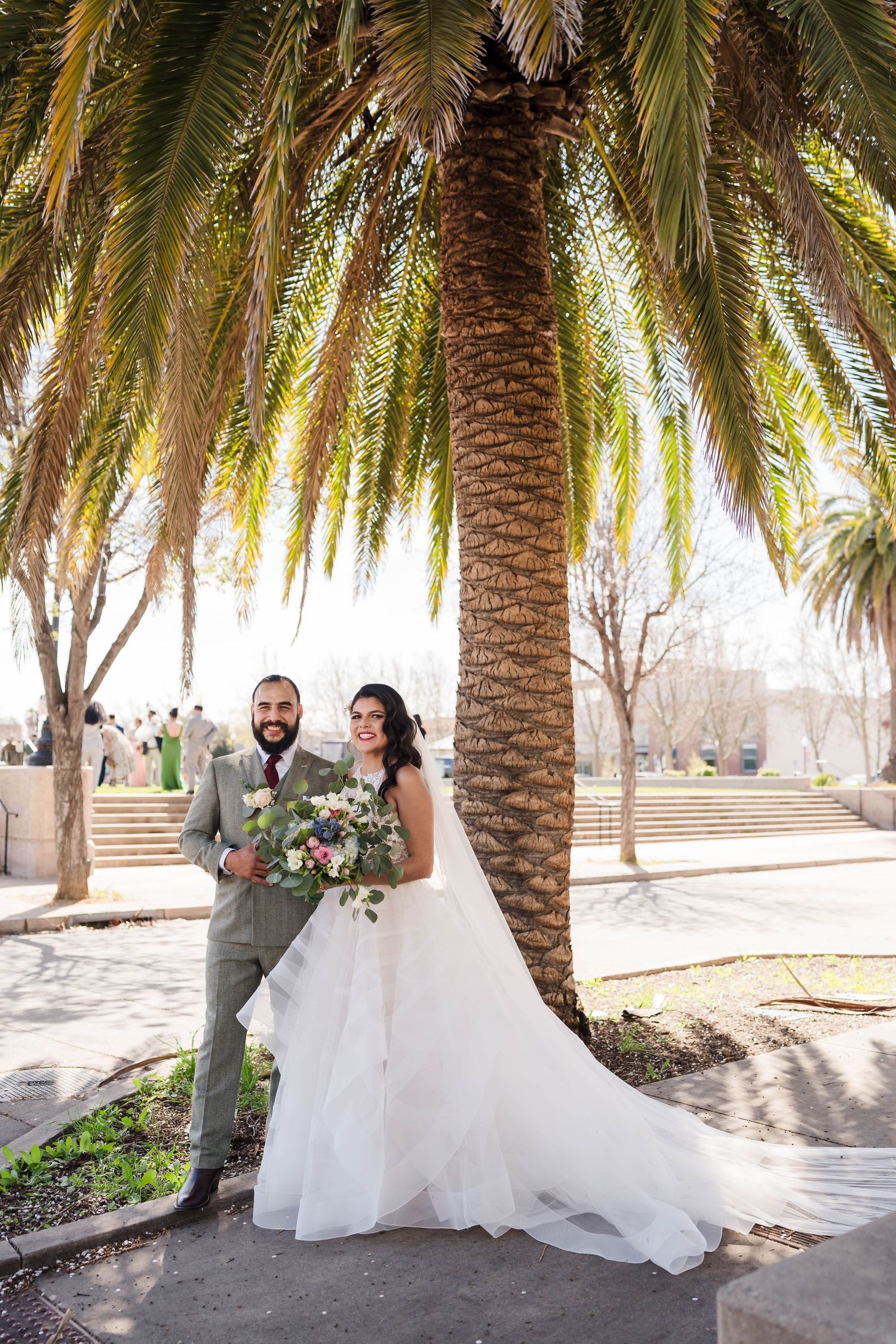 Bride and groom posing together under a large palm tree outdoors on their wedding day.