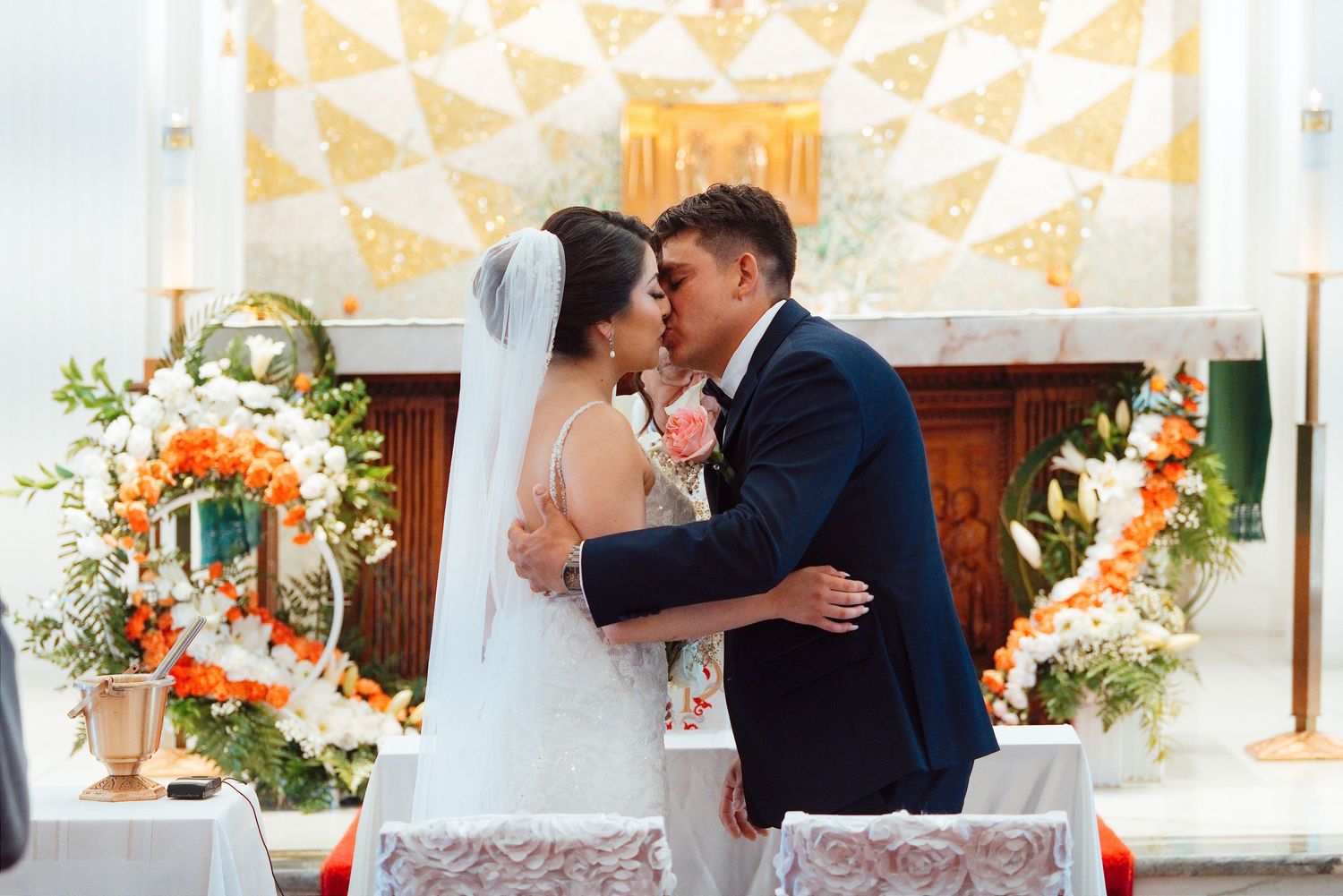 Bride and groom share first kiss at church altar surrounded by elegant orange and white floral arrangements.