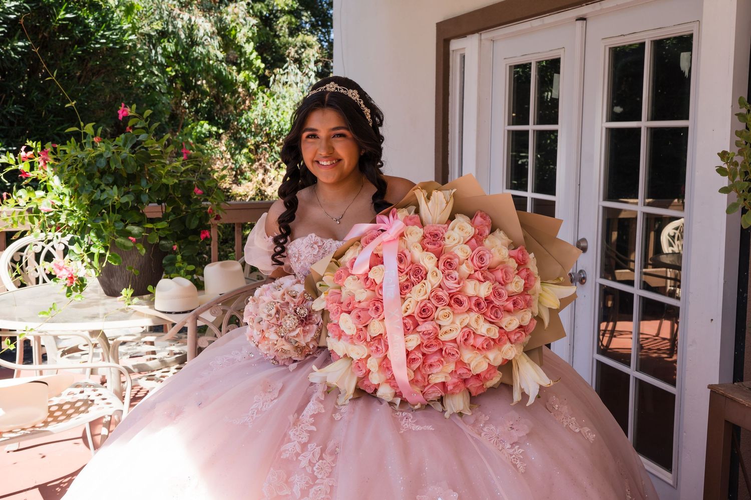 Young woman in pink quinceañera ball gown holding large bouquet of pink and white roses outdoors.