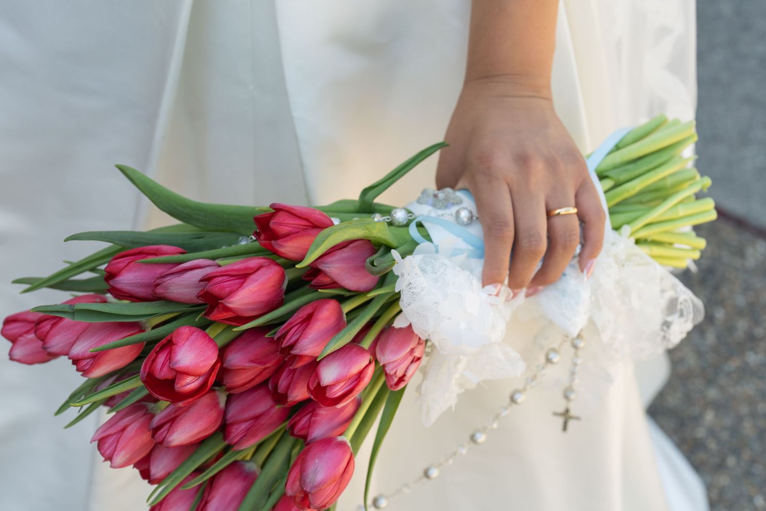 Bride holding a bouquet of red tulips with white lace and a rosary, wearing a gold wedding ring.