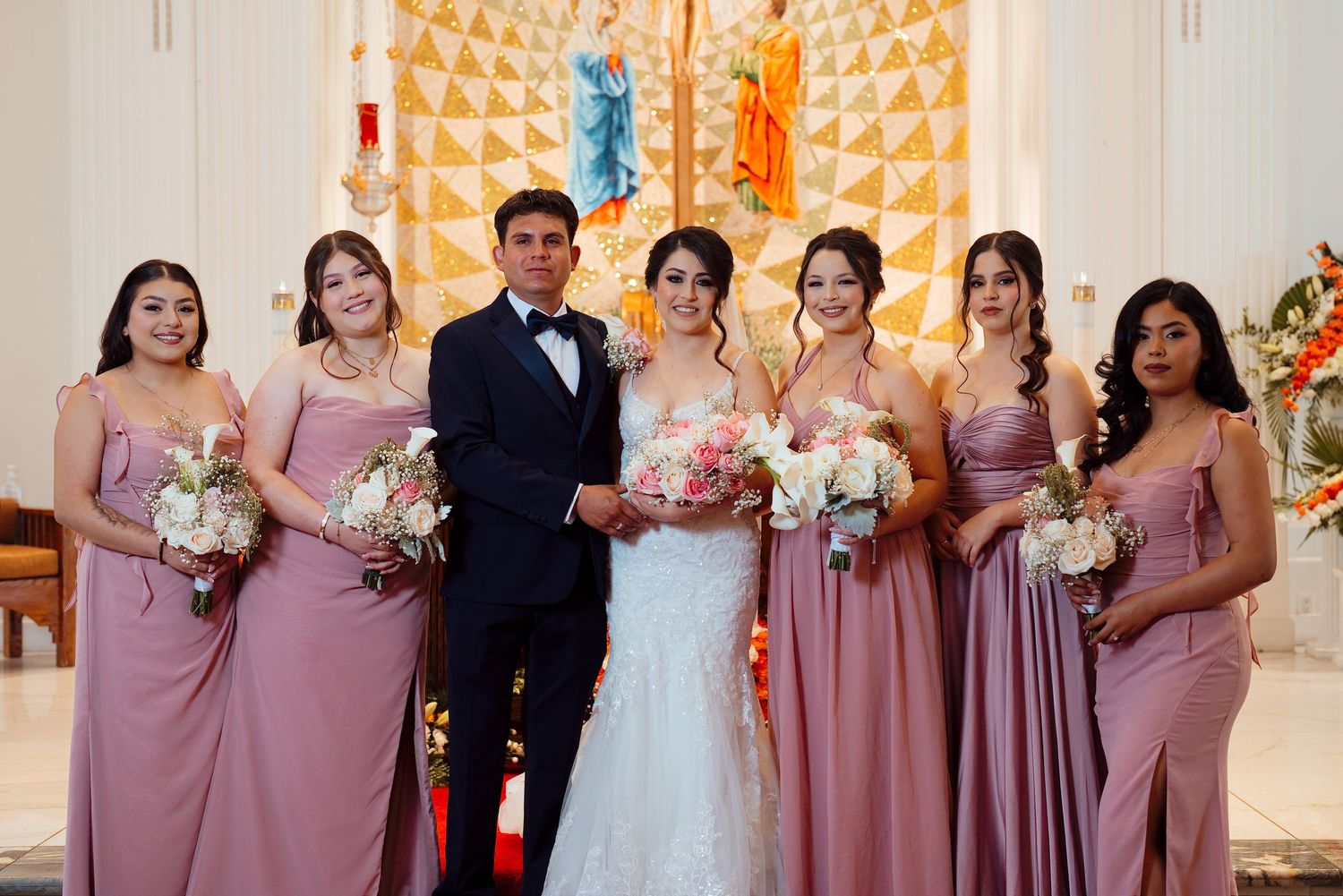 Bride and groom pose with bridesmaids in dusty rose gowns holding bouquets inside an elegant church.
