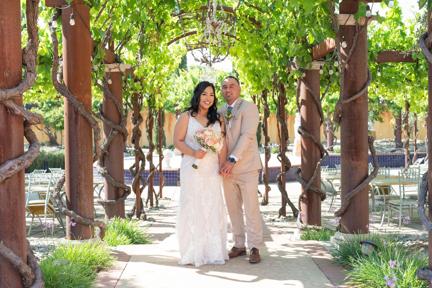 Bride and groom posing under a rustic vine-covered pergola at an outdoor wedding venue on a sunny day.