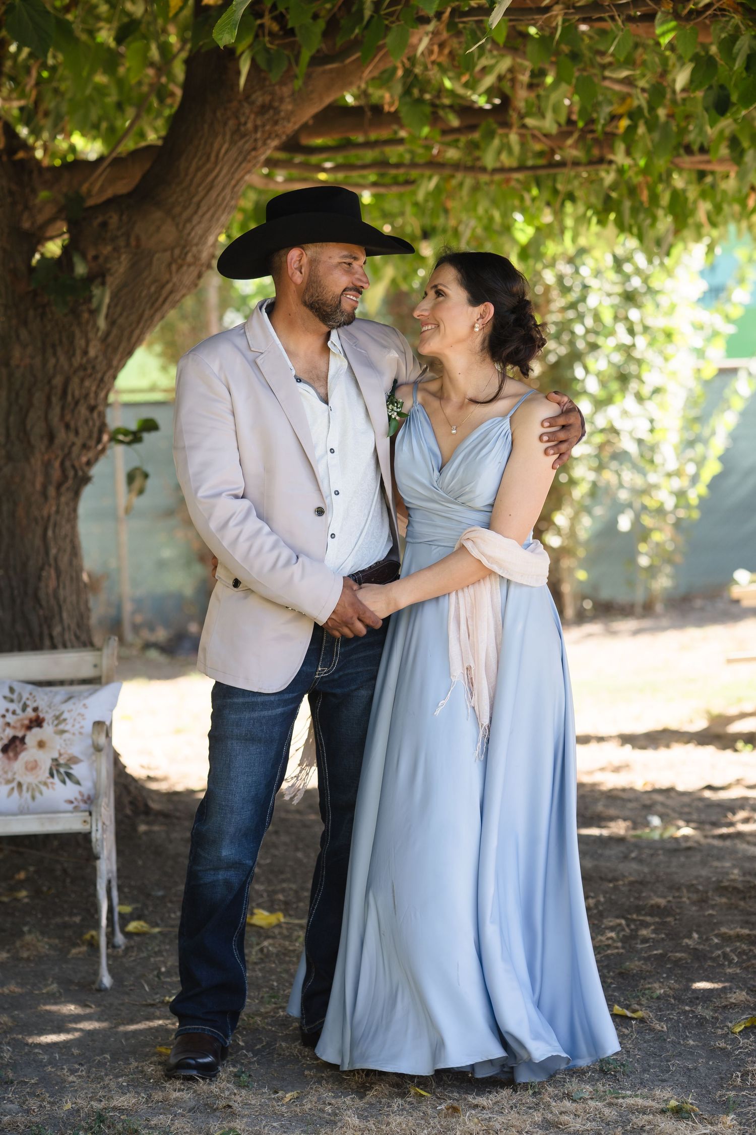 Couple in rustic wedding attire embrace under a tree, man in cowboy hat and jeans, woman in blue gown.