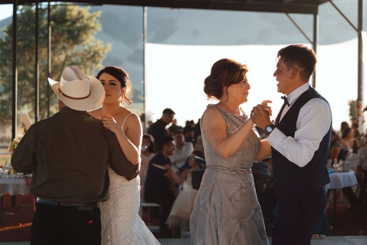 Two couples share parent dances at outdoor wedding reception with mountain views and guests in background.