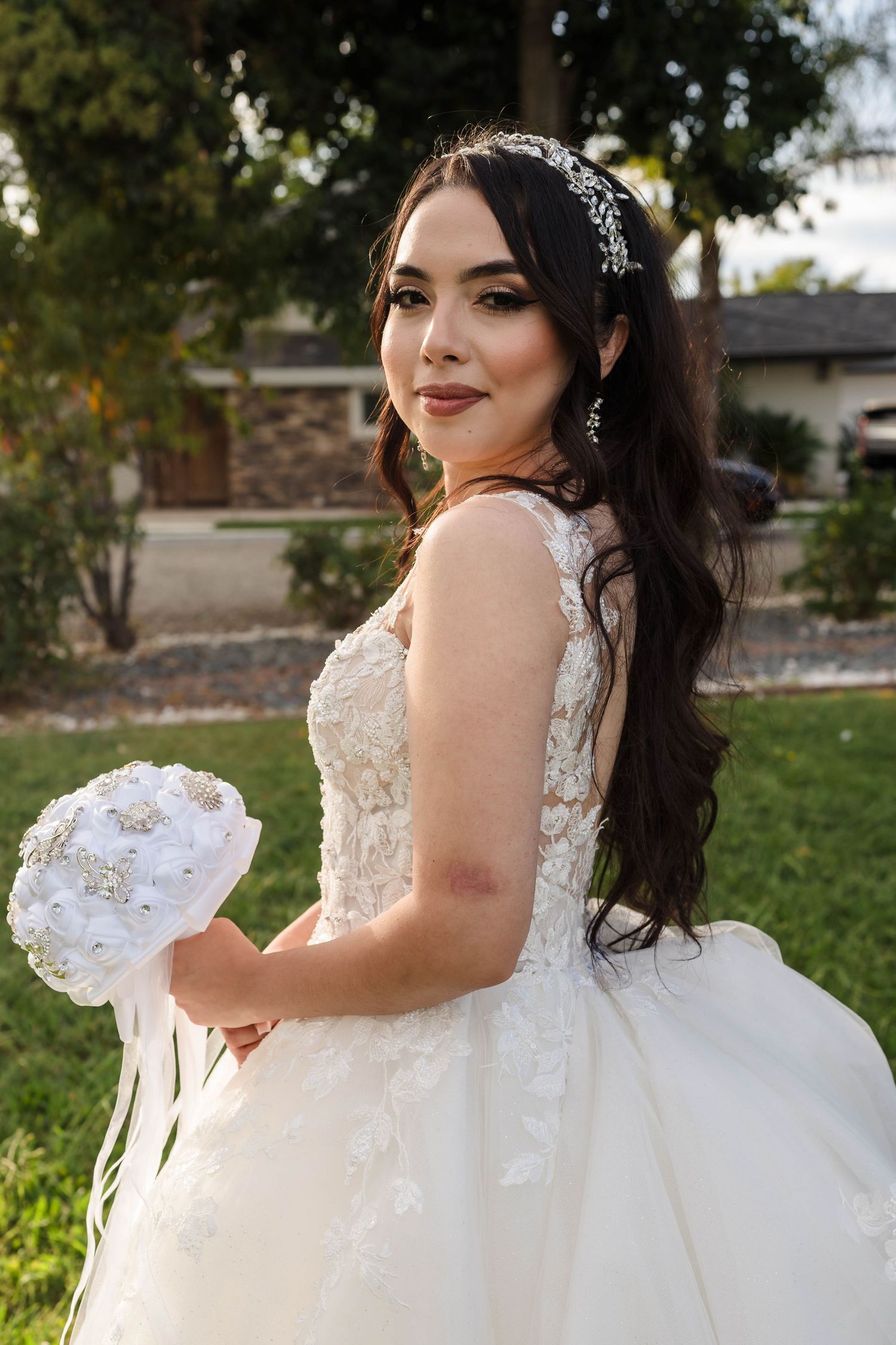Bride in white lace ballgown holding pearl brooch bouquet, wearing crystal hair accessory, posing outdoors on green lawn.