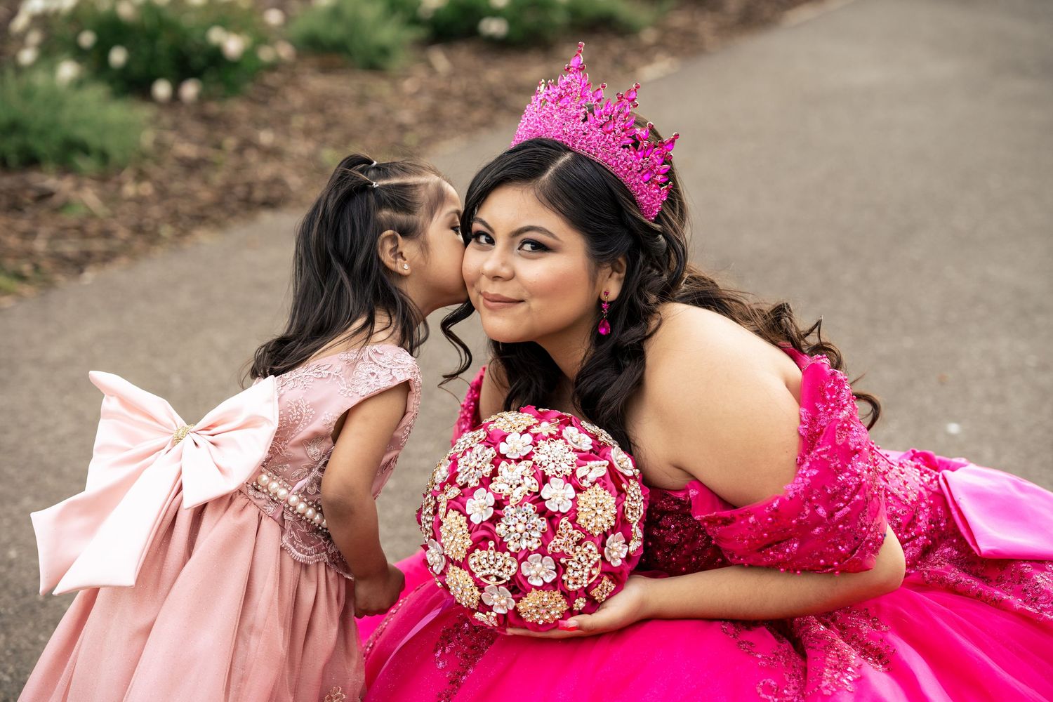 Quinceañera in pink crown holds jeweled bouquet as young girl in pink bow dress kisses her cheek.