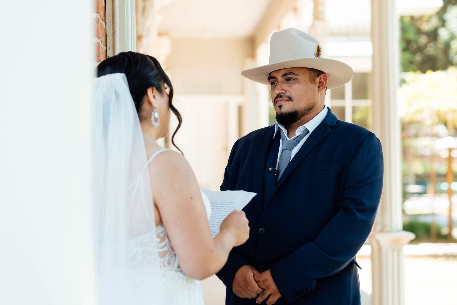 Groom in cowboy hat listens as bride reads wedding vows on a classic white-columned porch.