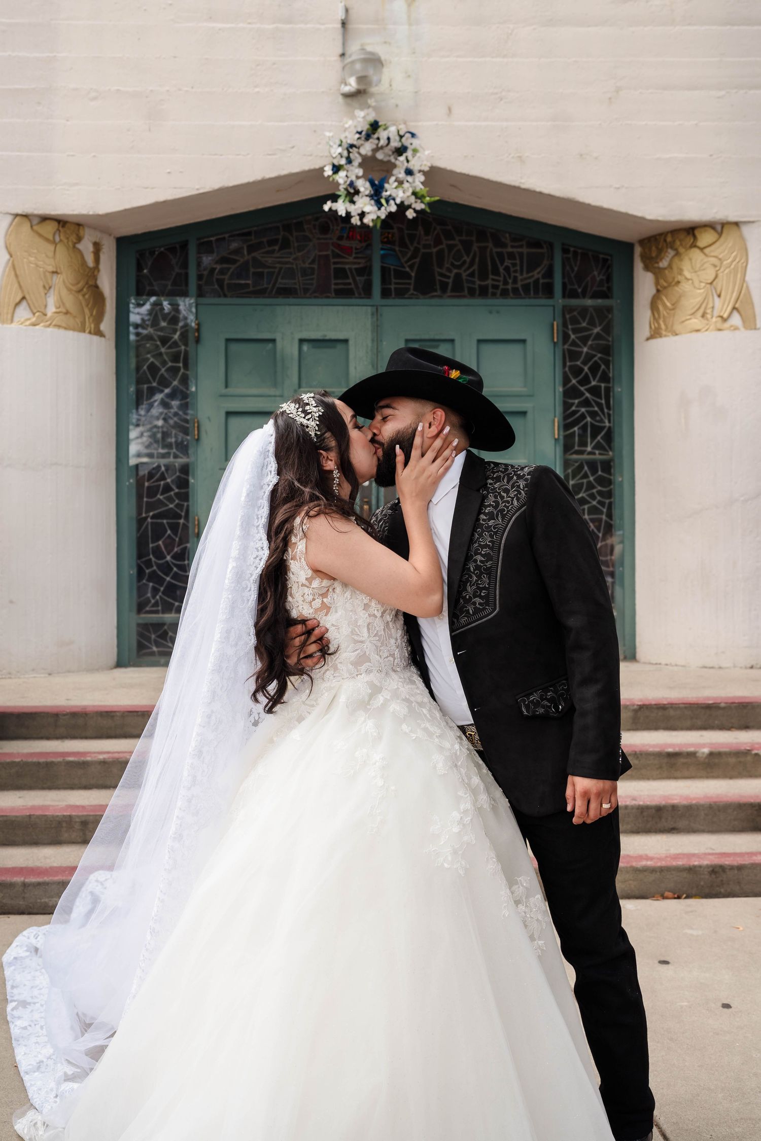 Bride and groom share a kiss in front of a church door, wearing a white ball gown and black charro suit with cowboy hat.