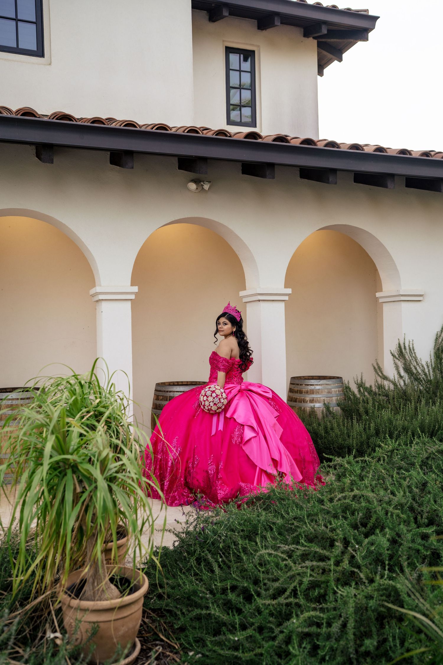 Young woman in voluminous hot pink quinceañera gown poses under white arched colonnade with barrels.