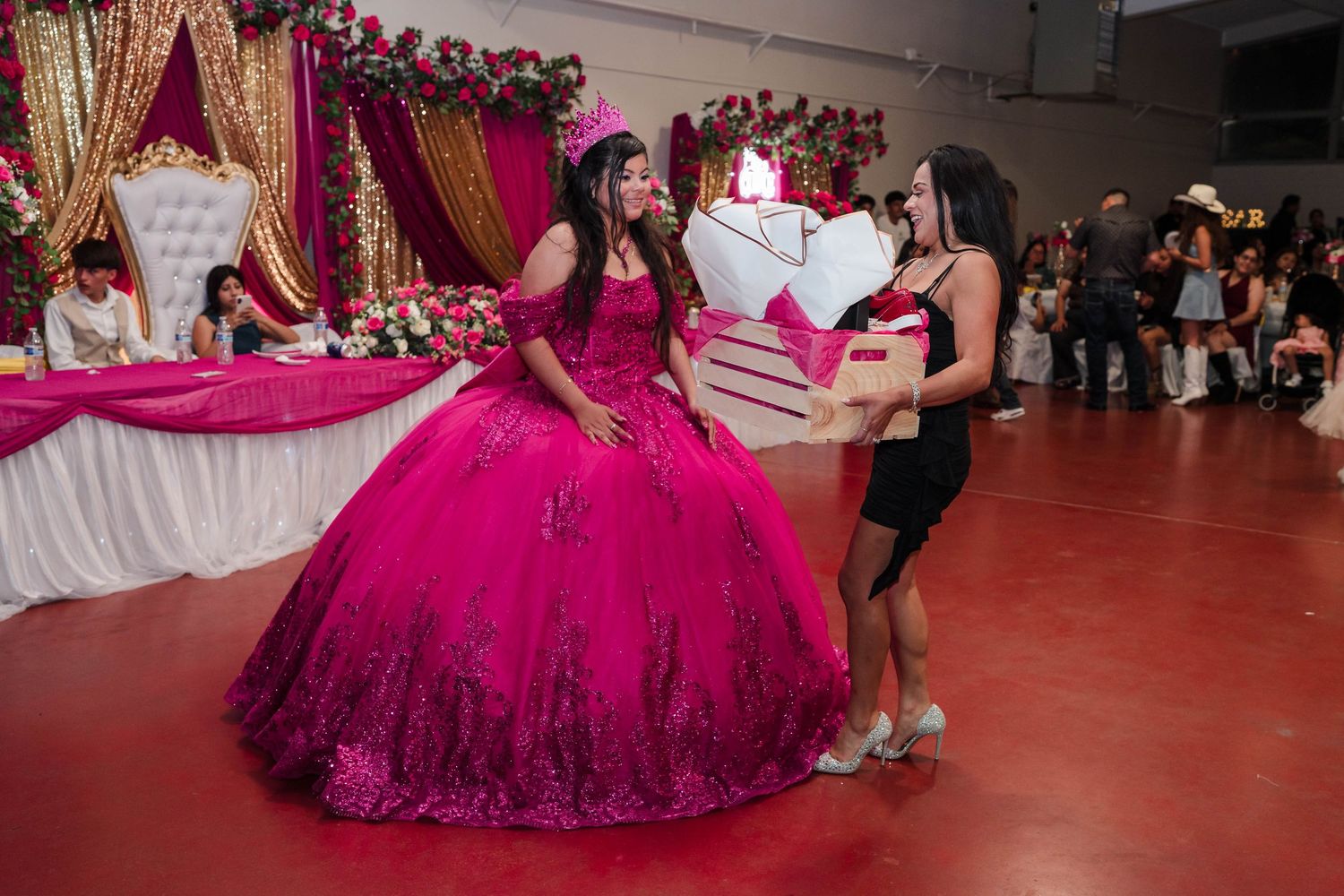 Quinceañera girl in a pink ball gown receives a heart-shaped gift box from a guest at her party.