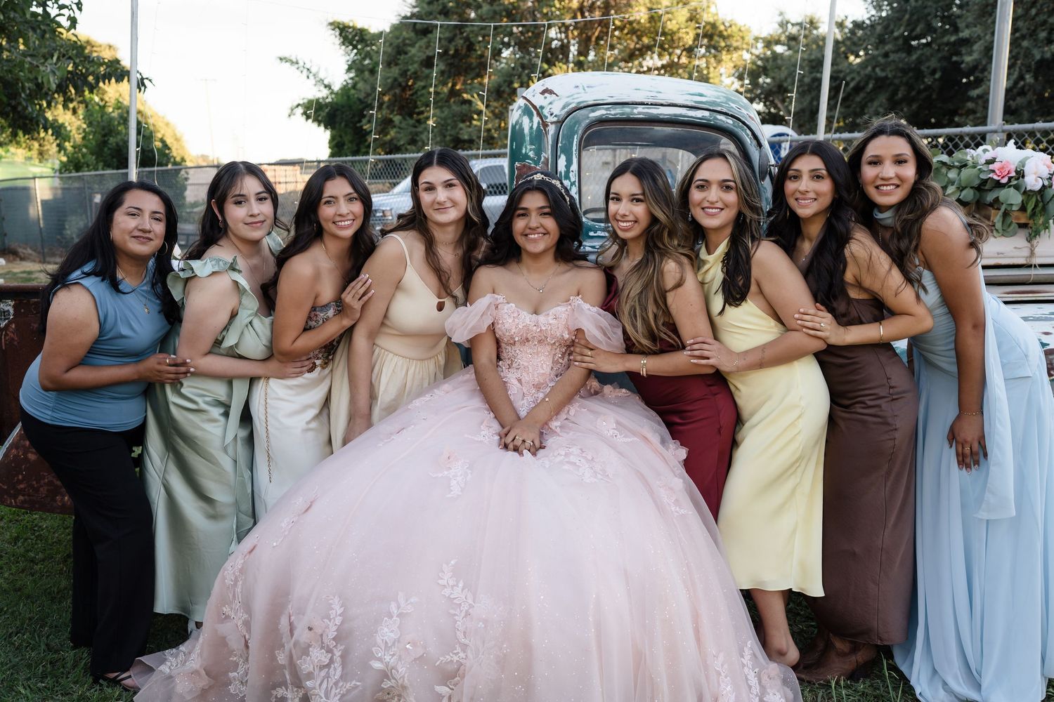 A young woman in a pink quinceañera ball gown poses with eight friends in formal dresses outdoors.