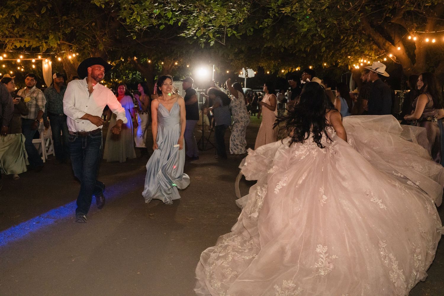 Guests dance outdoors at a quinceañera celebration under string lights with a girl in a pink gown.