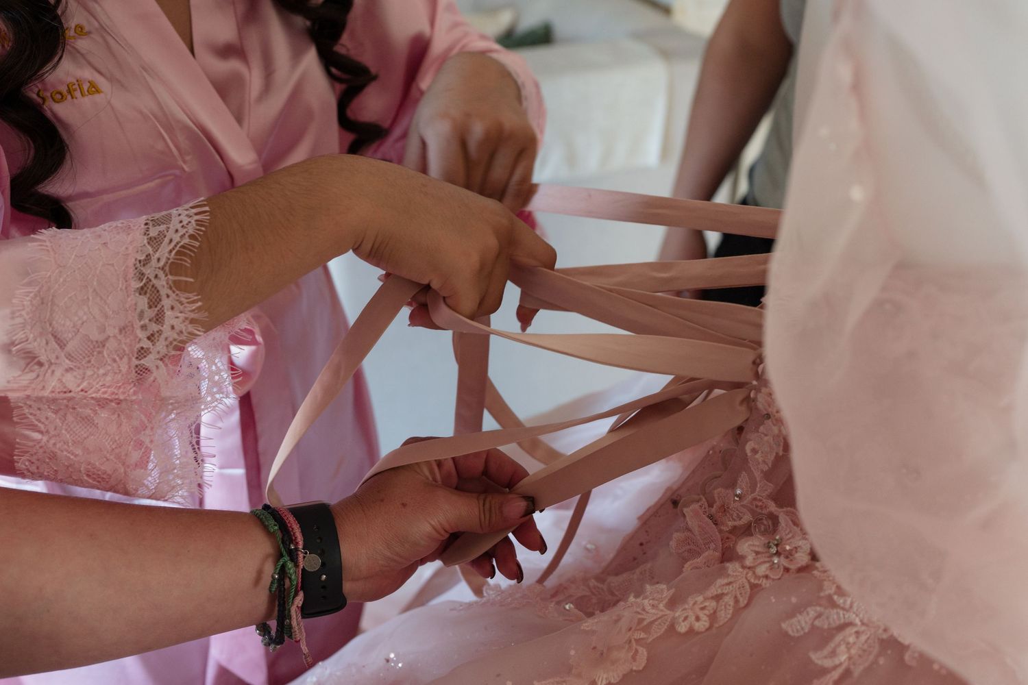 Bridesmaids in pink robes lacing up a bridal gown with satin ribbons during wedding preparations.
