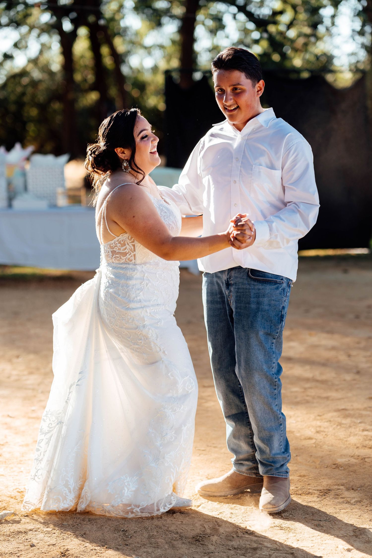 Bride in white lace gown and groom in white shirt and jeans share a dance in golden sunlight.