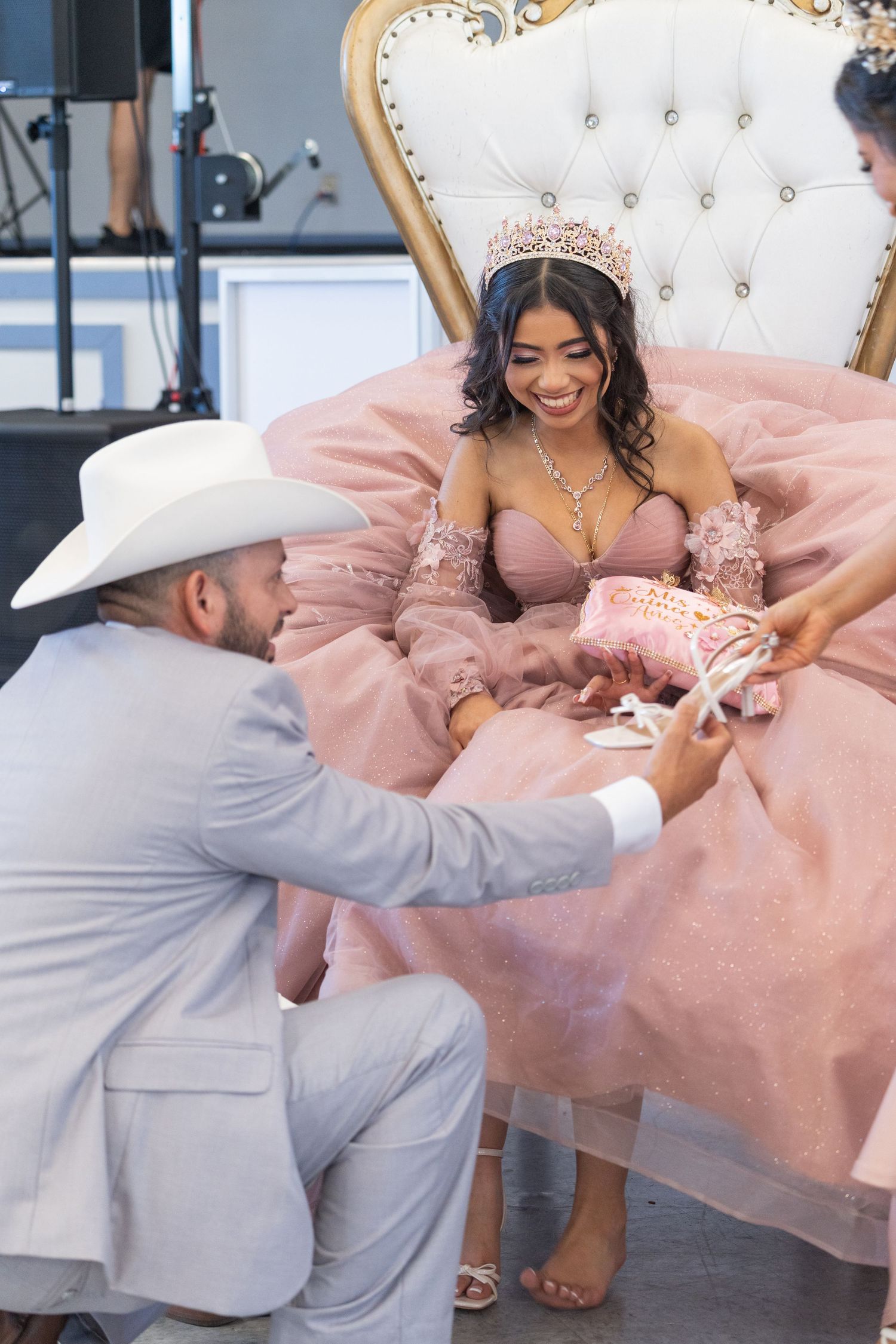 Smiling quinceañera girl in pink gown and tiara seated on ornate throne while man presents her with pink shoes.