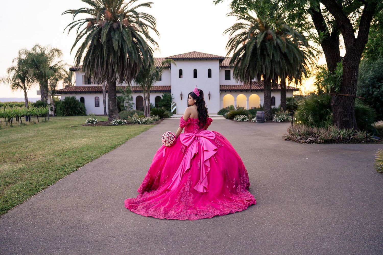 Quinceañera in stunning fuchsia ball gown walks path before elegant Spanish-style mansion at sunset.