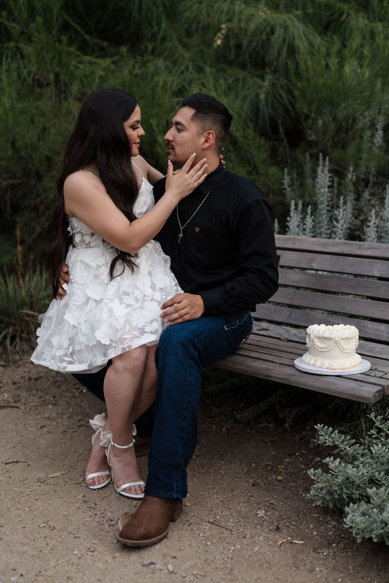 Couple sharing a romantic moment on a wooden bench with a white cake nearby in a lush garden setting.