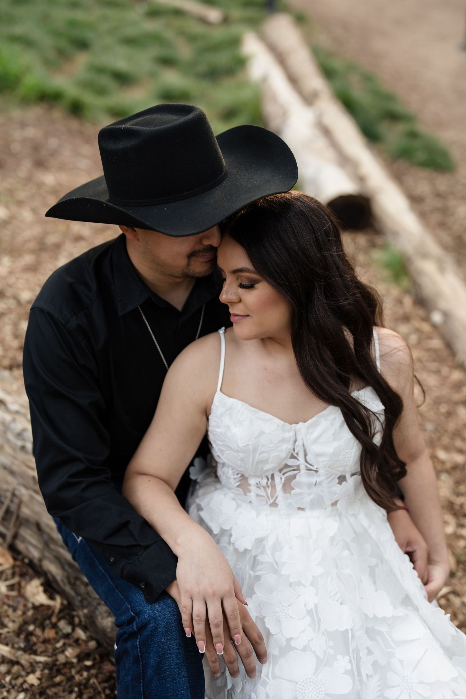 Engaged couple sitting on a log, woman in white floral dress leaning into man wearing black cowboy hat.