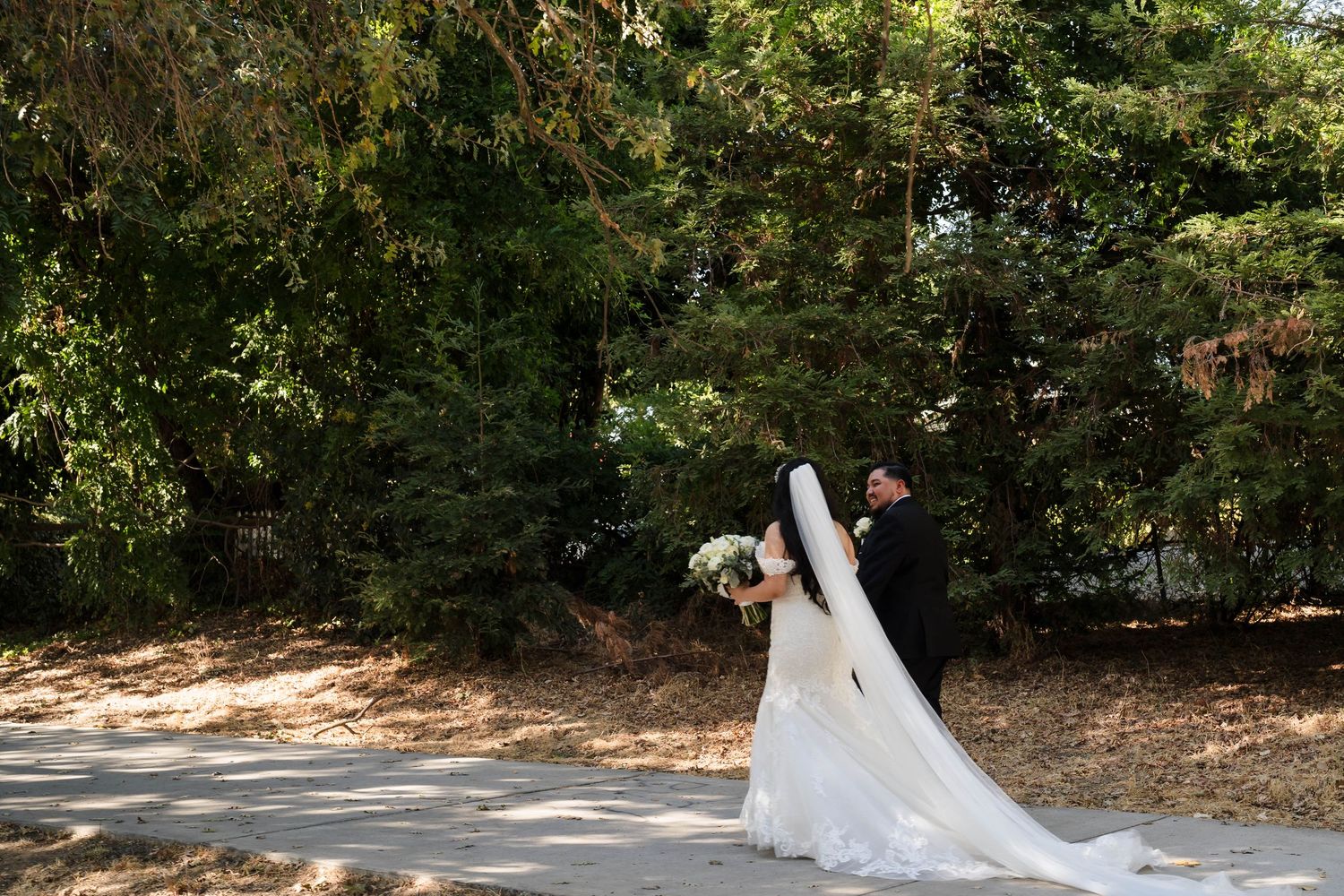 Bride in elegant white gown with long train holds bouquet while embracing groom near tall green trees.
