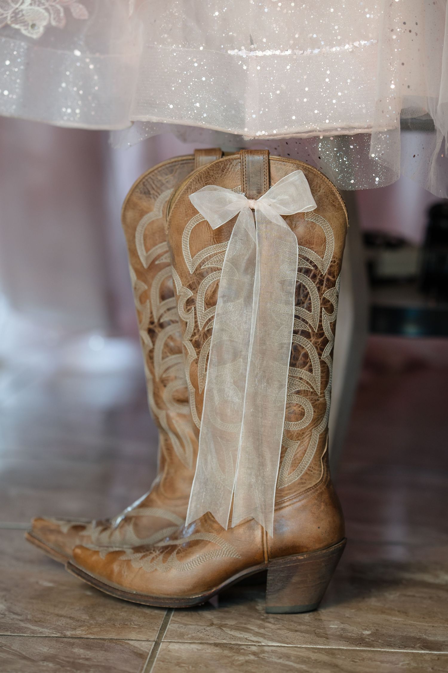 Tan cowboy boots with intricate stitching and a white ribbon bow, worn under a sparkly white dress on a tile floor.