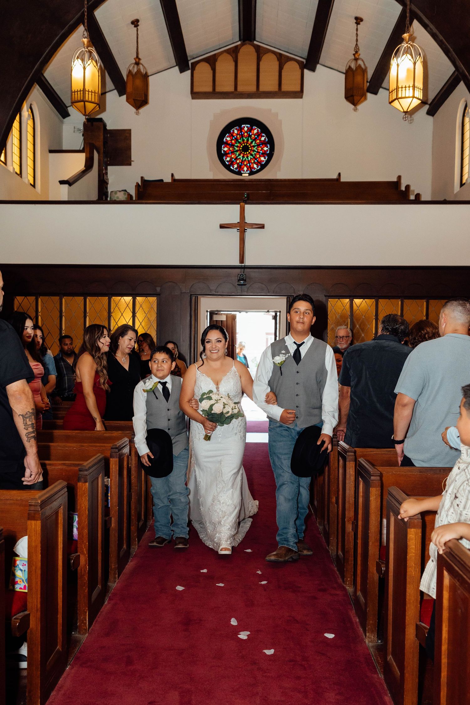Bride walks down red-carpeted church aisle with escorts during wedding ceremony.