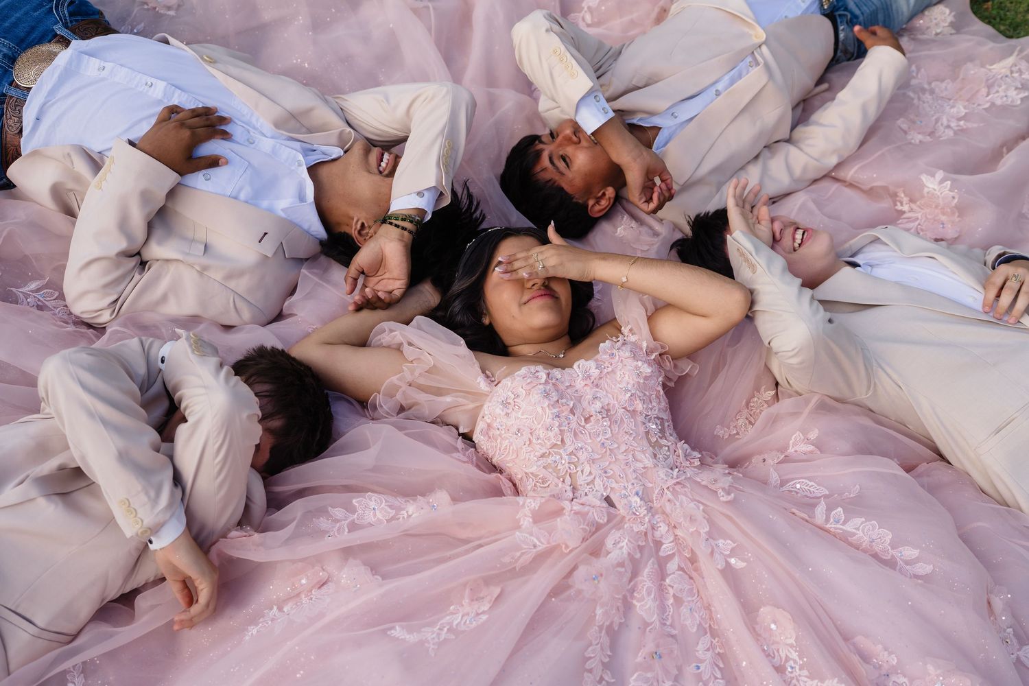 Quinceañera girl in pink ball gown lies on grass surrounded by chambelanes in a candid celebration photo.
