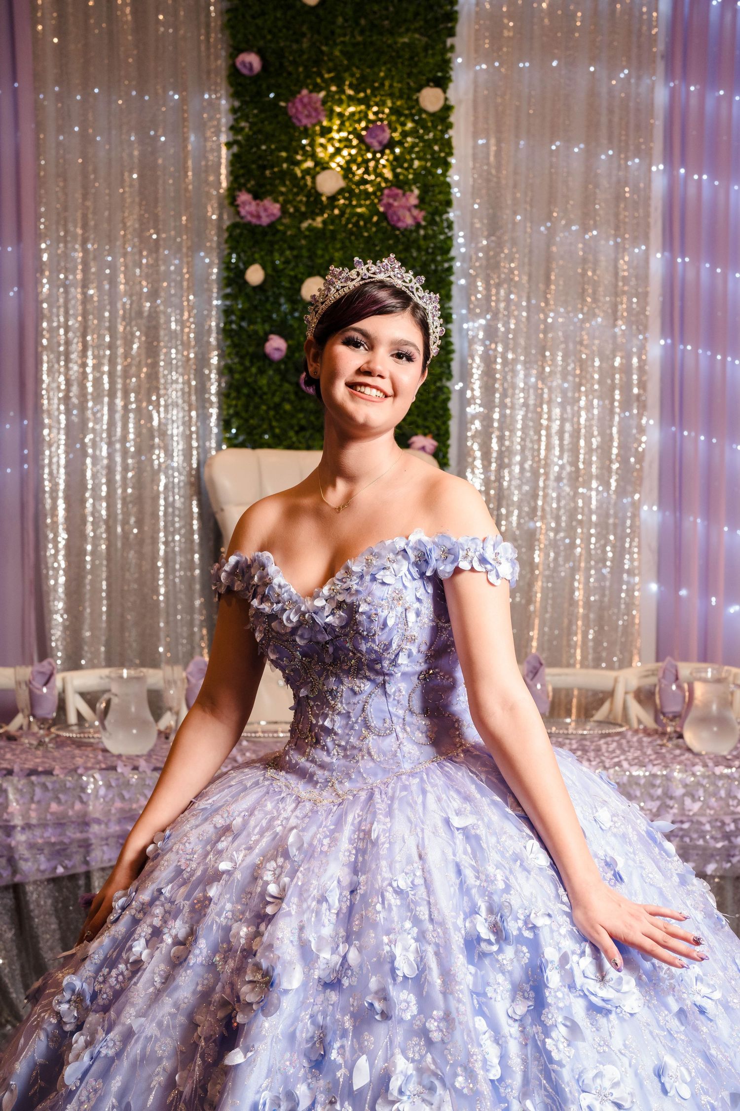 Smiling young woman in lavender ball gown with floral details and tiara at a quinceañera celebration.
