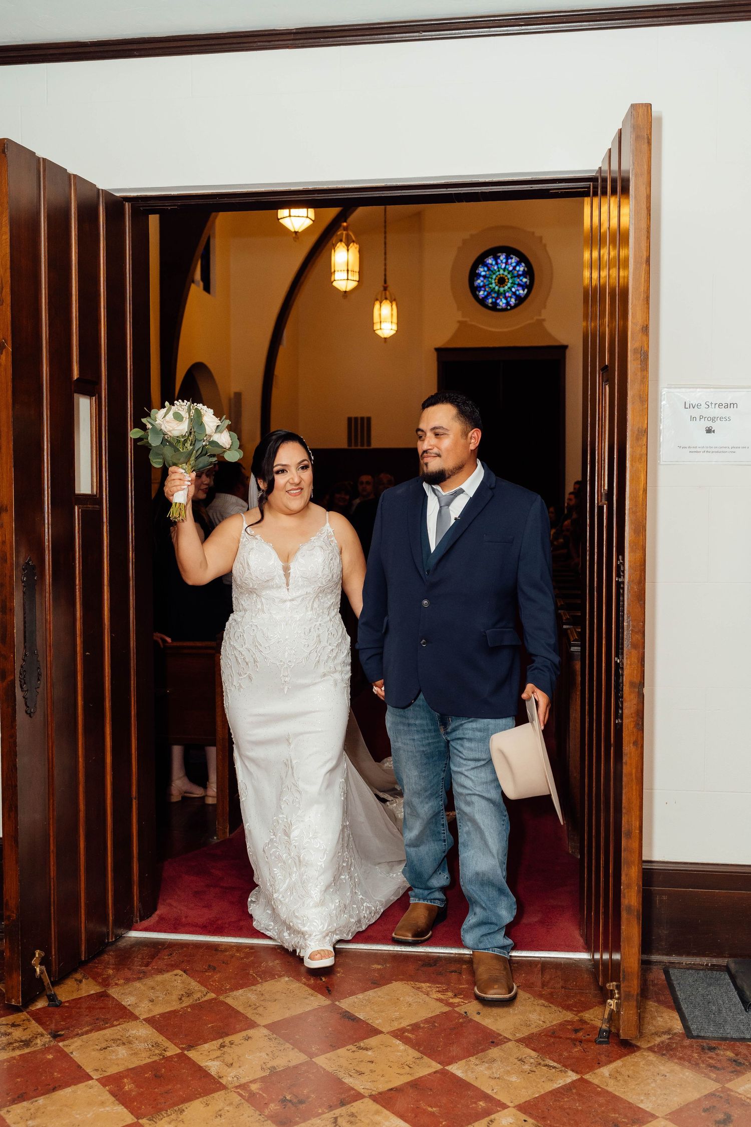 Newlyweds exit church doors smiling, bride raising bouquet in celebration.
