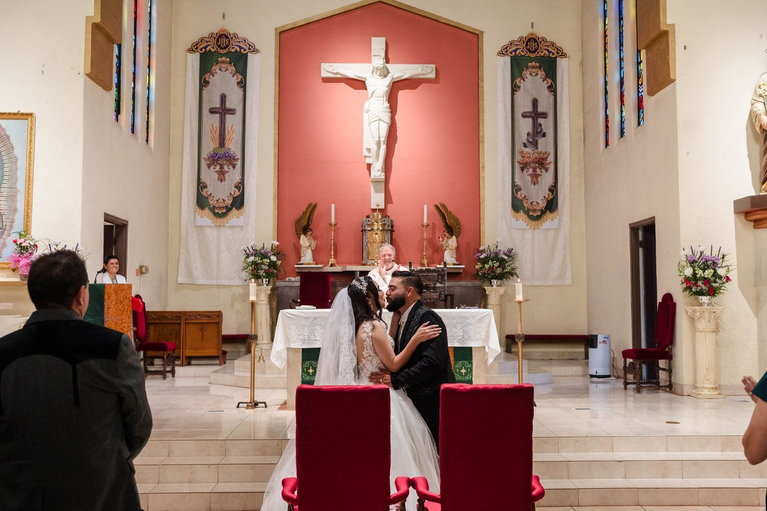 Bride and groom share a kiss at the altar inside a beautiful Catholic church decorated with flowers and a crucifix.