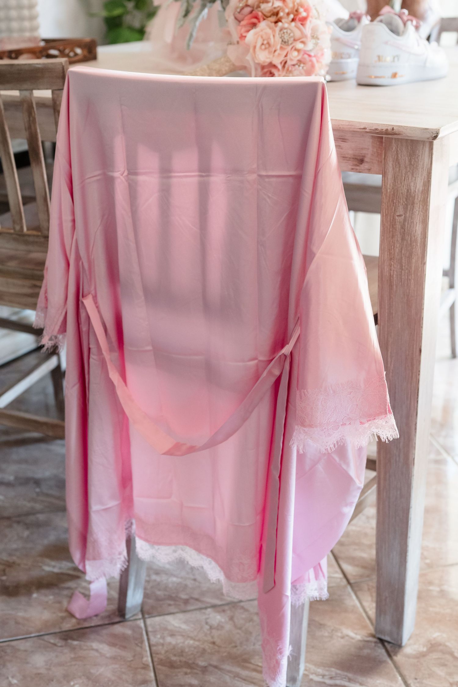 Pink lace-trimmed bridal robe draped over a wooden chair at a elegant wedding preparation setting.