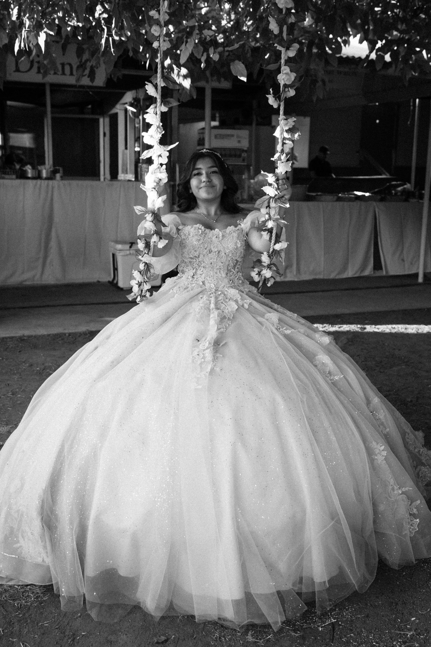 Young woman in white off-shoulder ballgown sitting on floral swing, black and white quinceañera portrait.