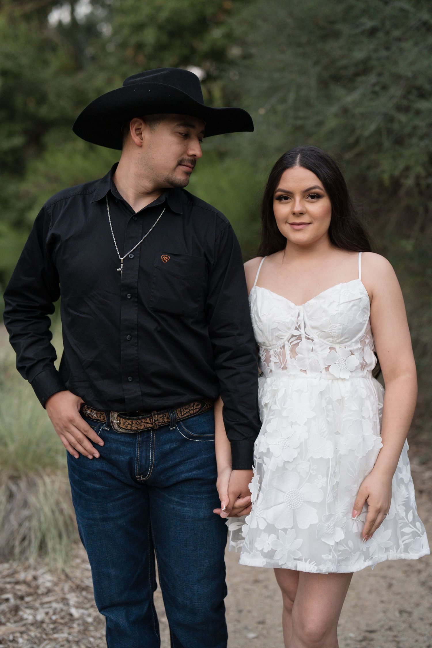 Couple holding hands outdoors, man in black cowboy hat and shirt, woman in white floral mini dress.