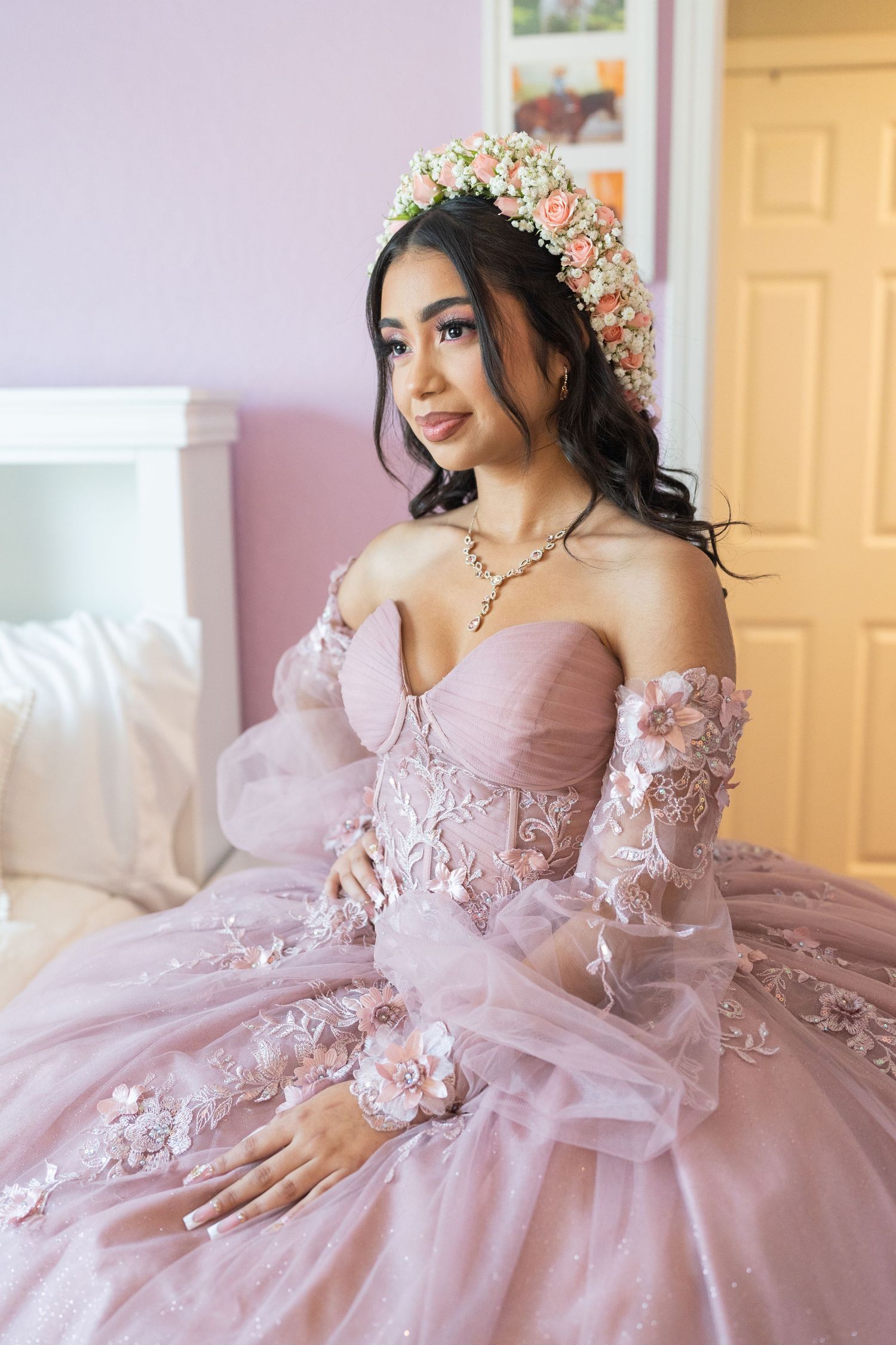 Young woman in a pink off-shoulder quinceañera gown with floral crown sitting gracefully on a bed.