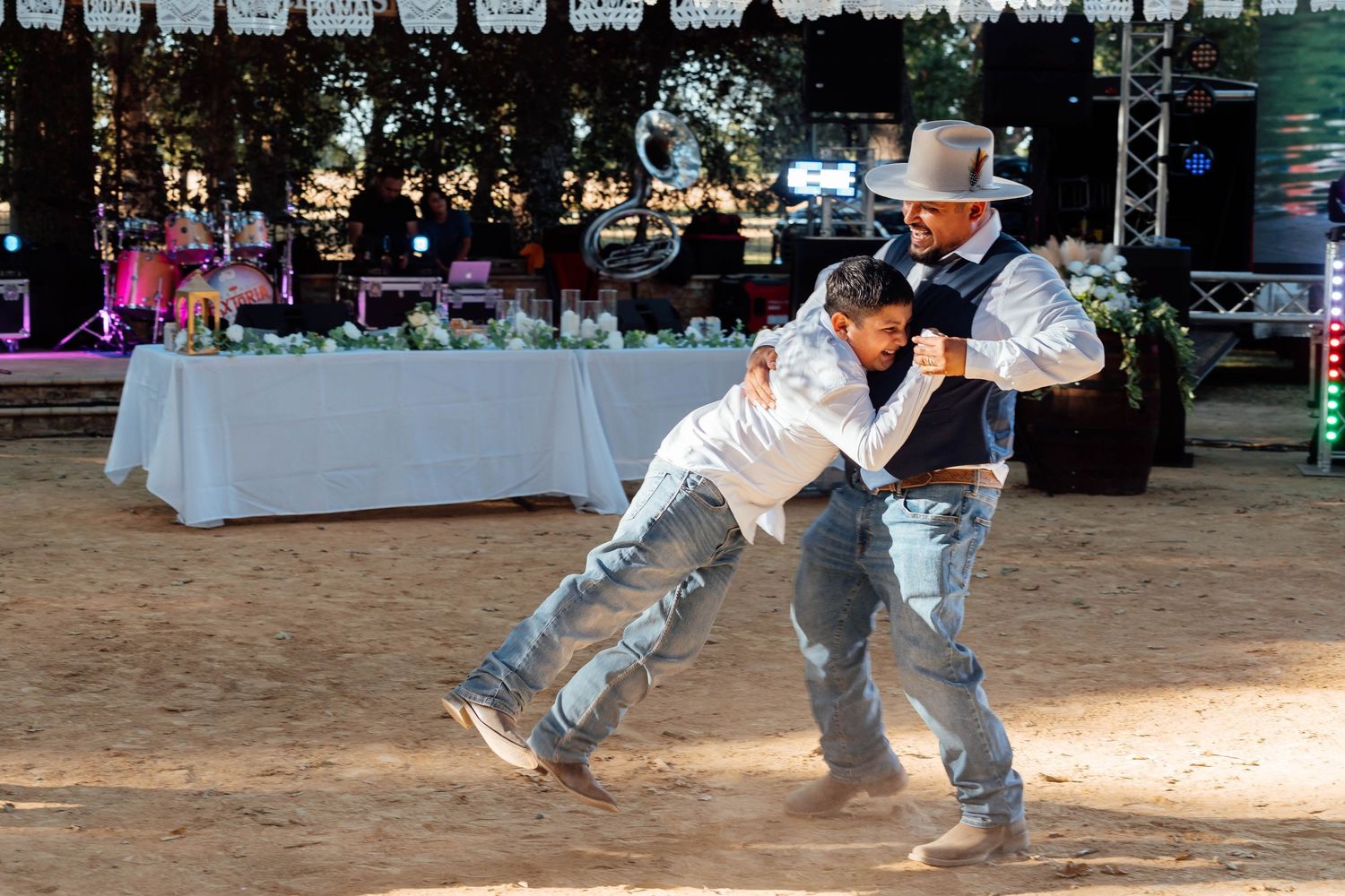 Two men playfully dance together at an outdoor rustic wedding reception near decorated tables.