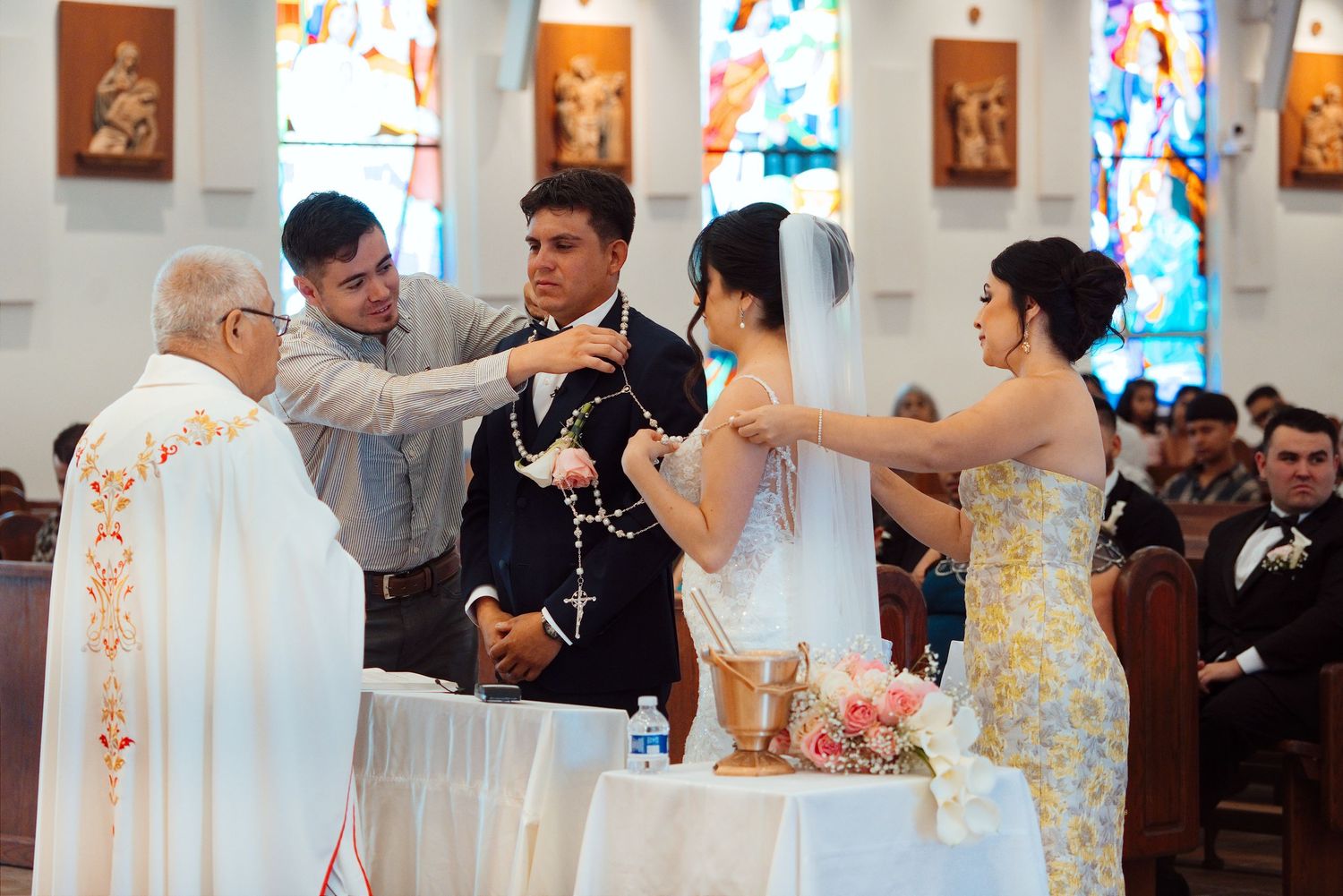 Catholic wedding ceremony with priest, couple, and wedding party exchanging rosary at church altar.