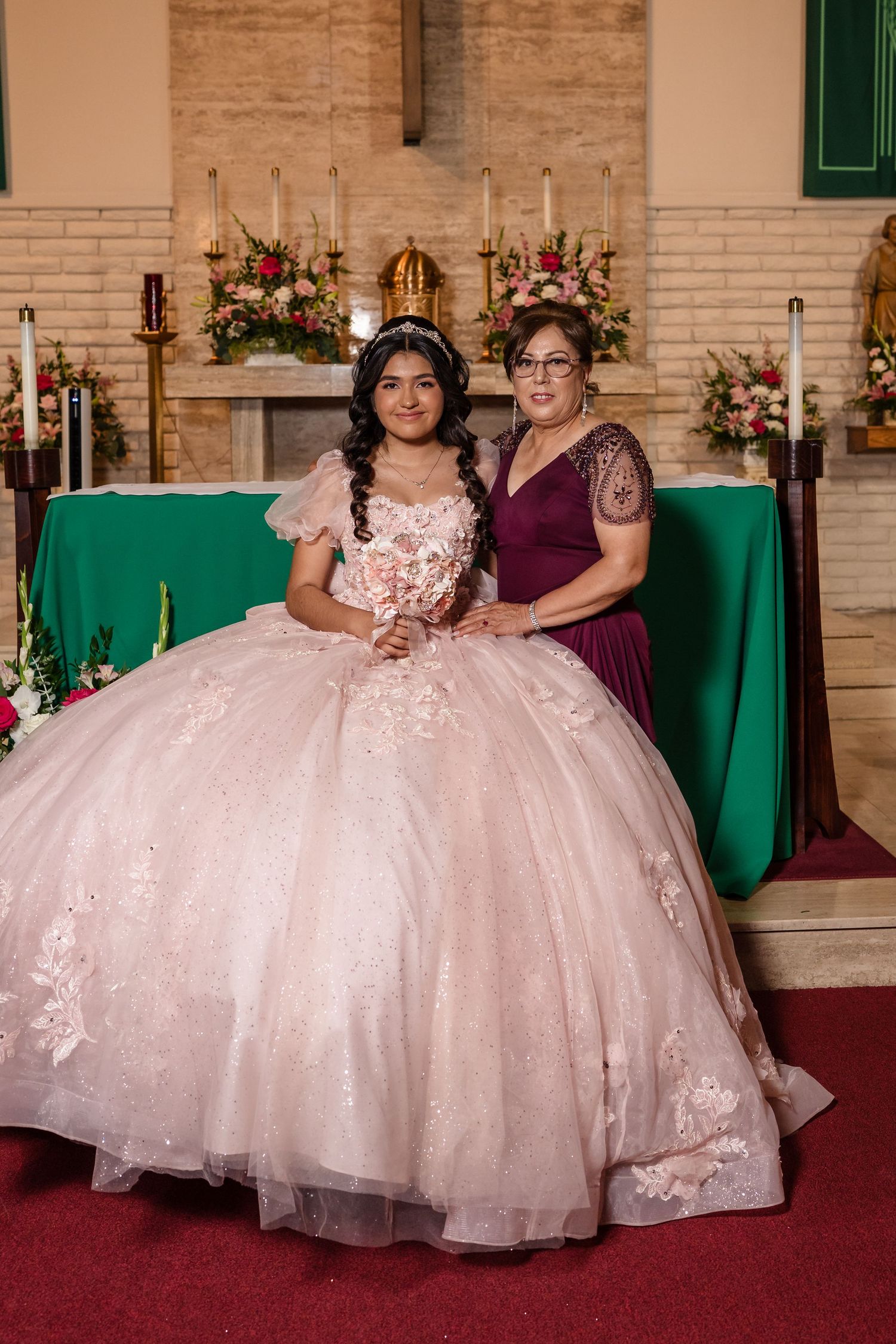Young woman in pink quinceañera ball gown poses with older woman in burgundy dress at church altar.