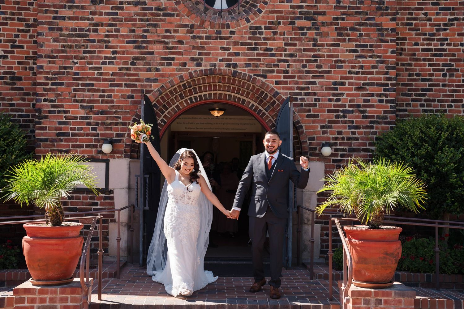 Newlywed couple celebrating outside a brick church, bride holding bouquet aloft while holding groom's hand on sunny wedding day.