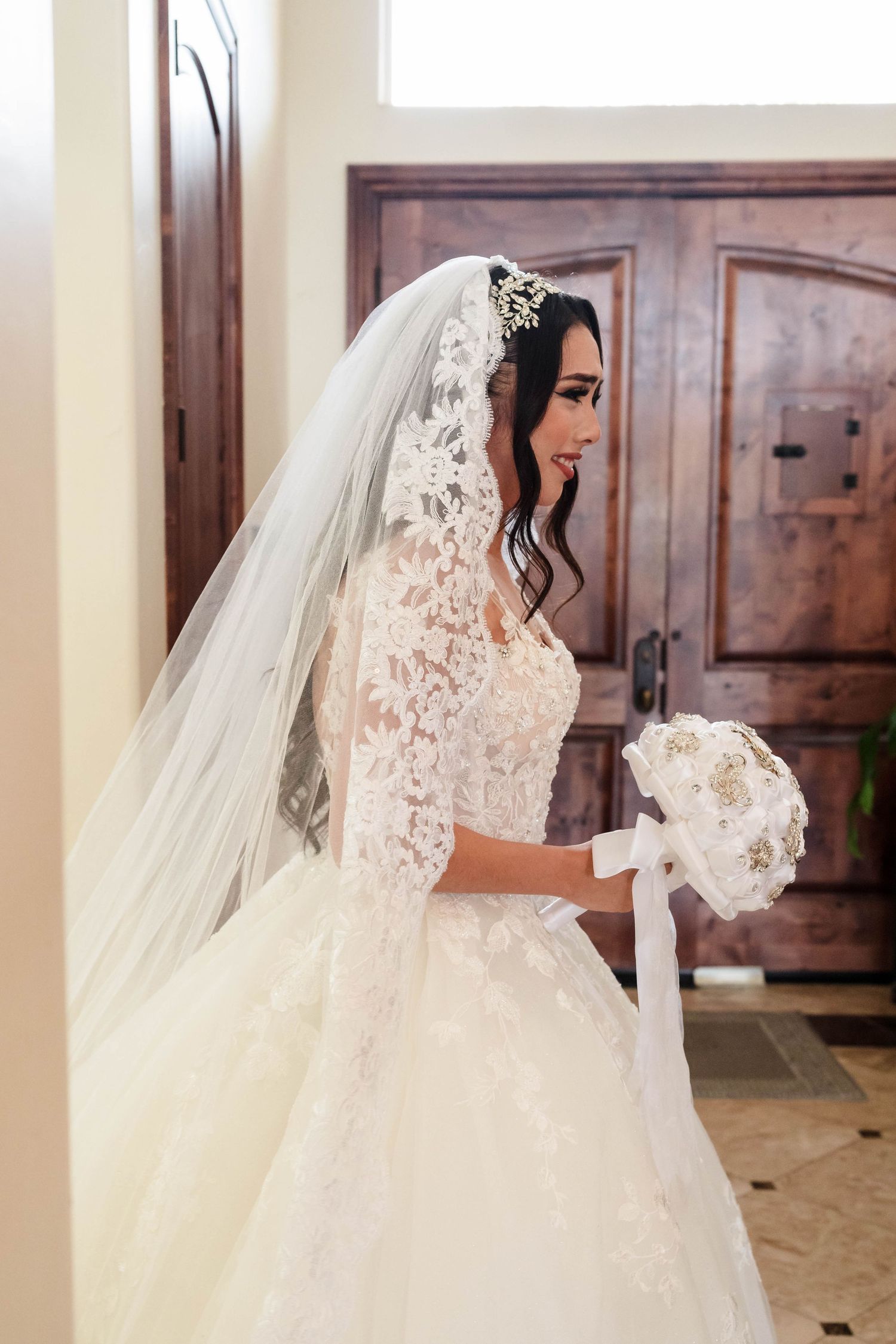 Bride in lace ball gown with cathedral veil and floral headpiece holds white bouquet near wooden doors.